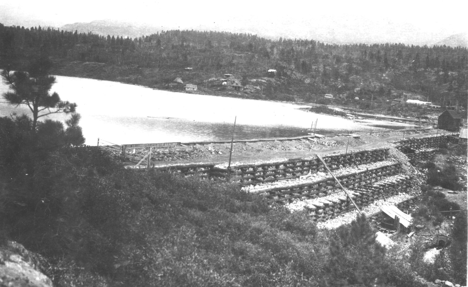 A reservoir held behind a long, low timber-faced embankment, with a small structure on the crest and buildings among trees on the far shore.