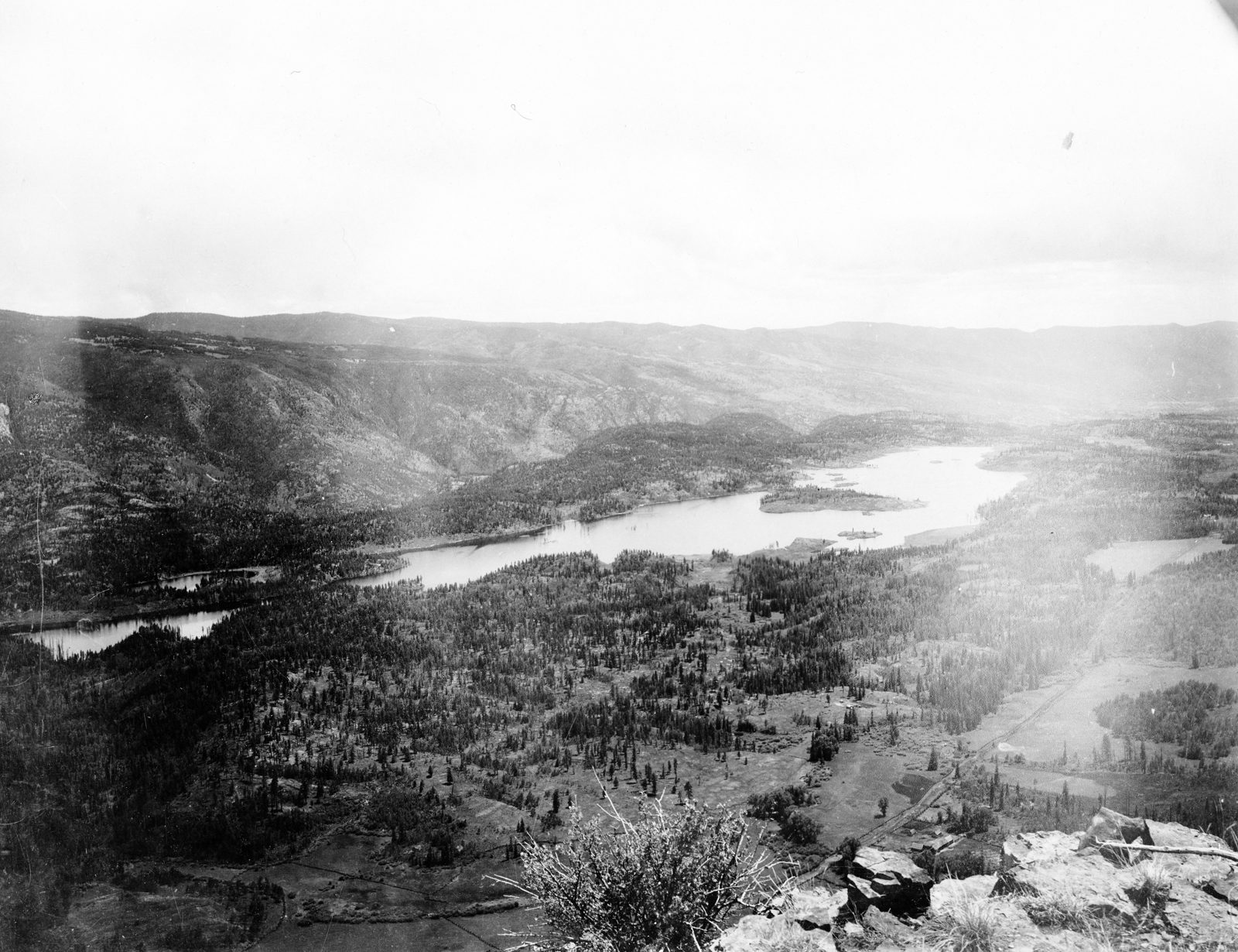 A long reservoir lies in a broad timbered valley seen from a clifftop, with cleared fields along the far shore and low ranges fading into haze beyond.