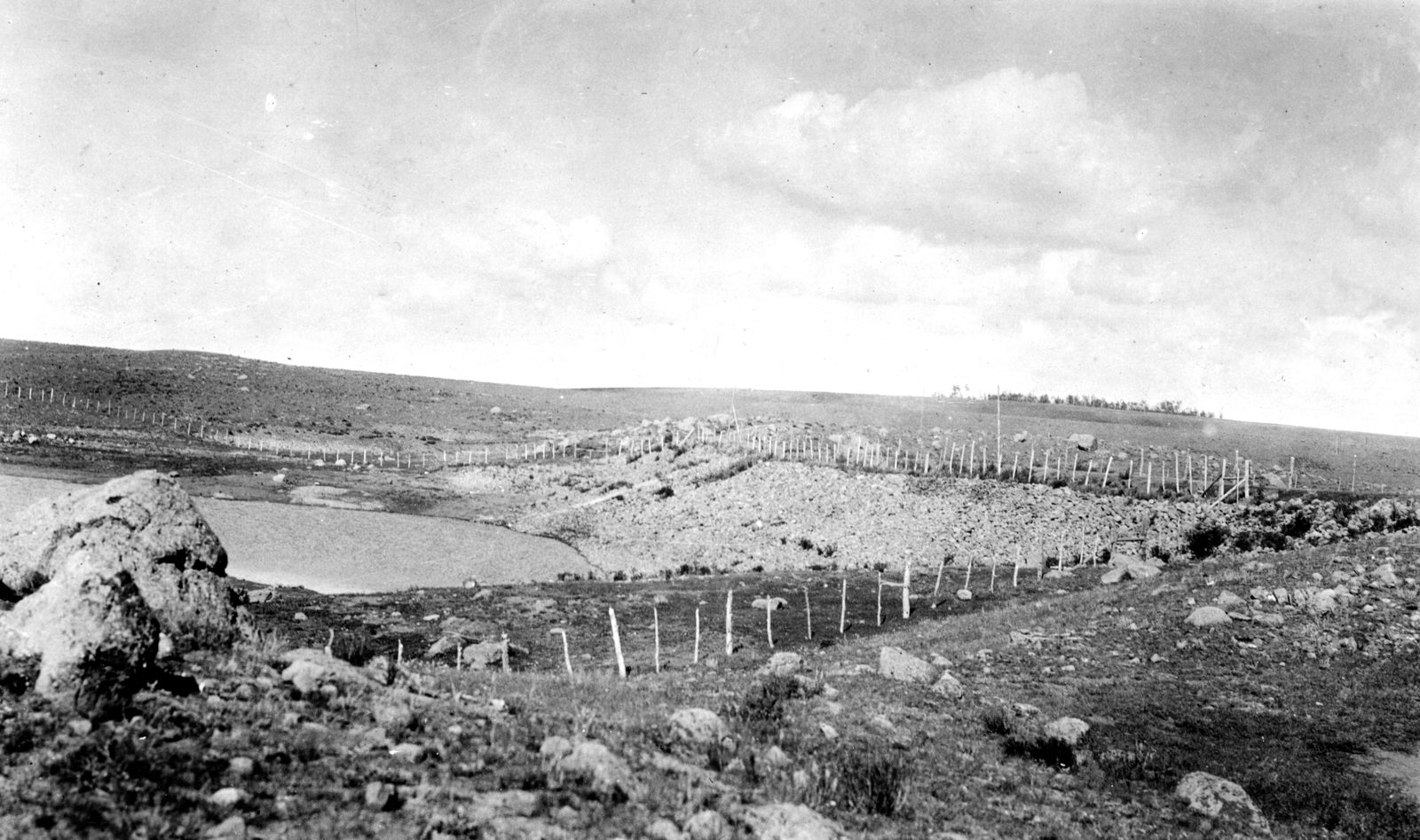 A low rock-fill dam of loose stone across a shallow draw on open, treeless high ground, water pooled behind it; a line of fence posts runs along the crest under a bright, cloudy sky.