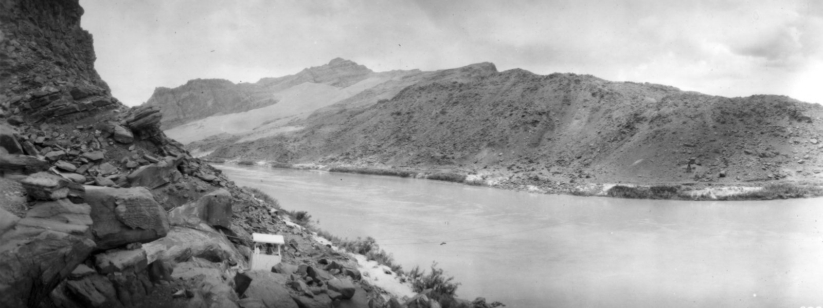 A wide silt-laden river runs between low desert bluffs, a small timber gage shelter standing on the near bank with a cable strung out across the water.