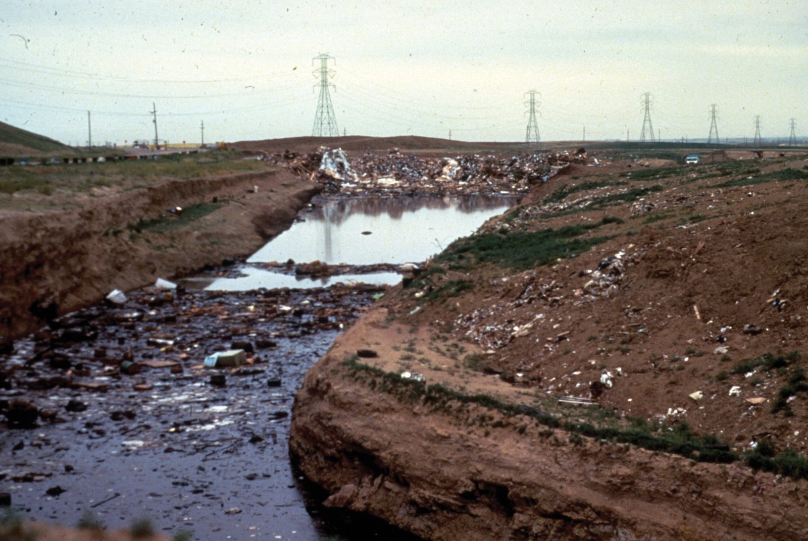 A shallow channel of dark standing water running between raw earth banks, refuse scattered along the right bank and across the fill behind, with transmission towers on the skyline.