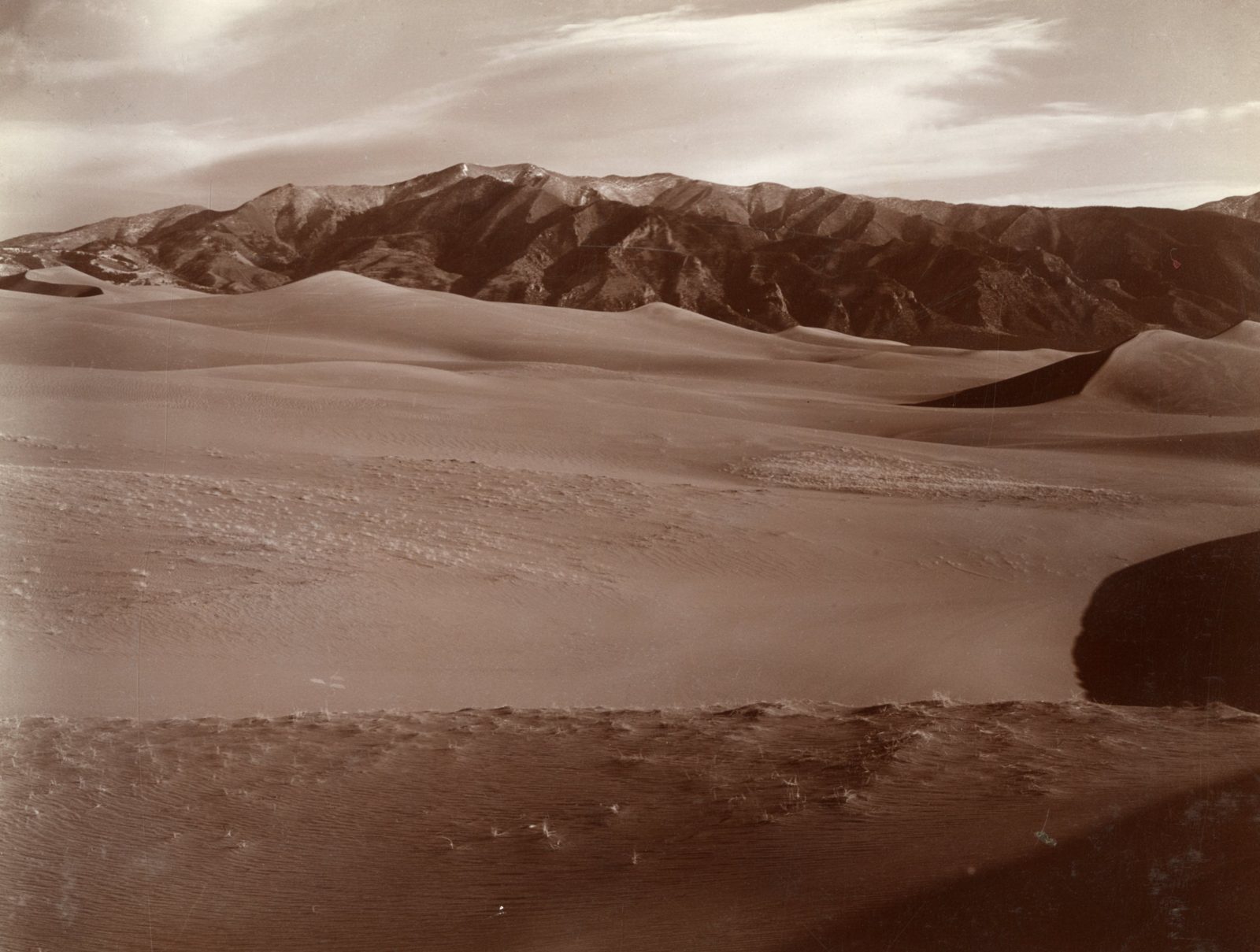 A long sand dune curving across a flat valley floor, its crest sharp against a low range of mountains behind, with loose rippled sand in the foreground under a pale sky.