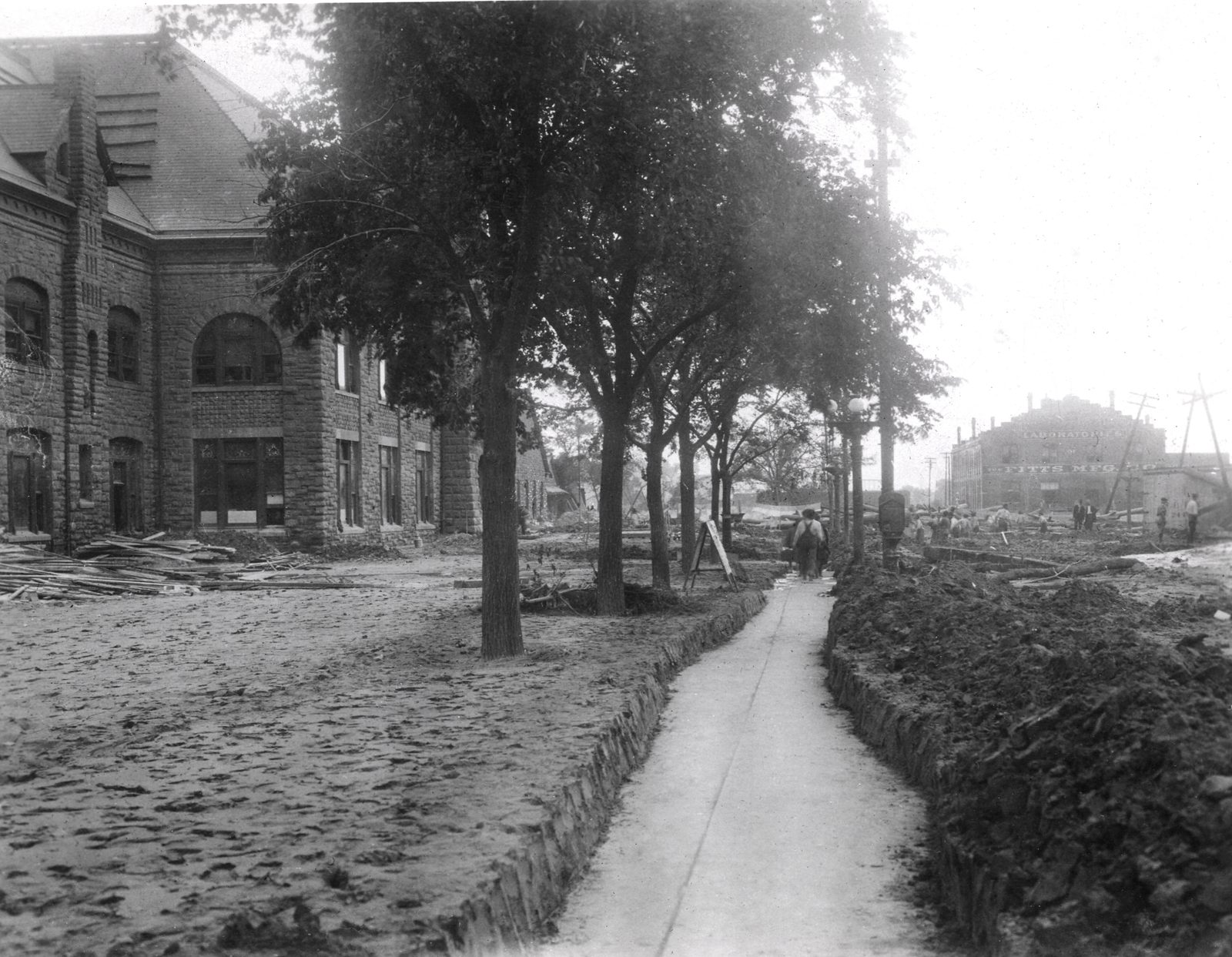 A cleared footpath cuts through a street buried in pale dried silt beside a stone depot, the mud banked in drifts around the tree trunks and men working in the distance.