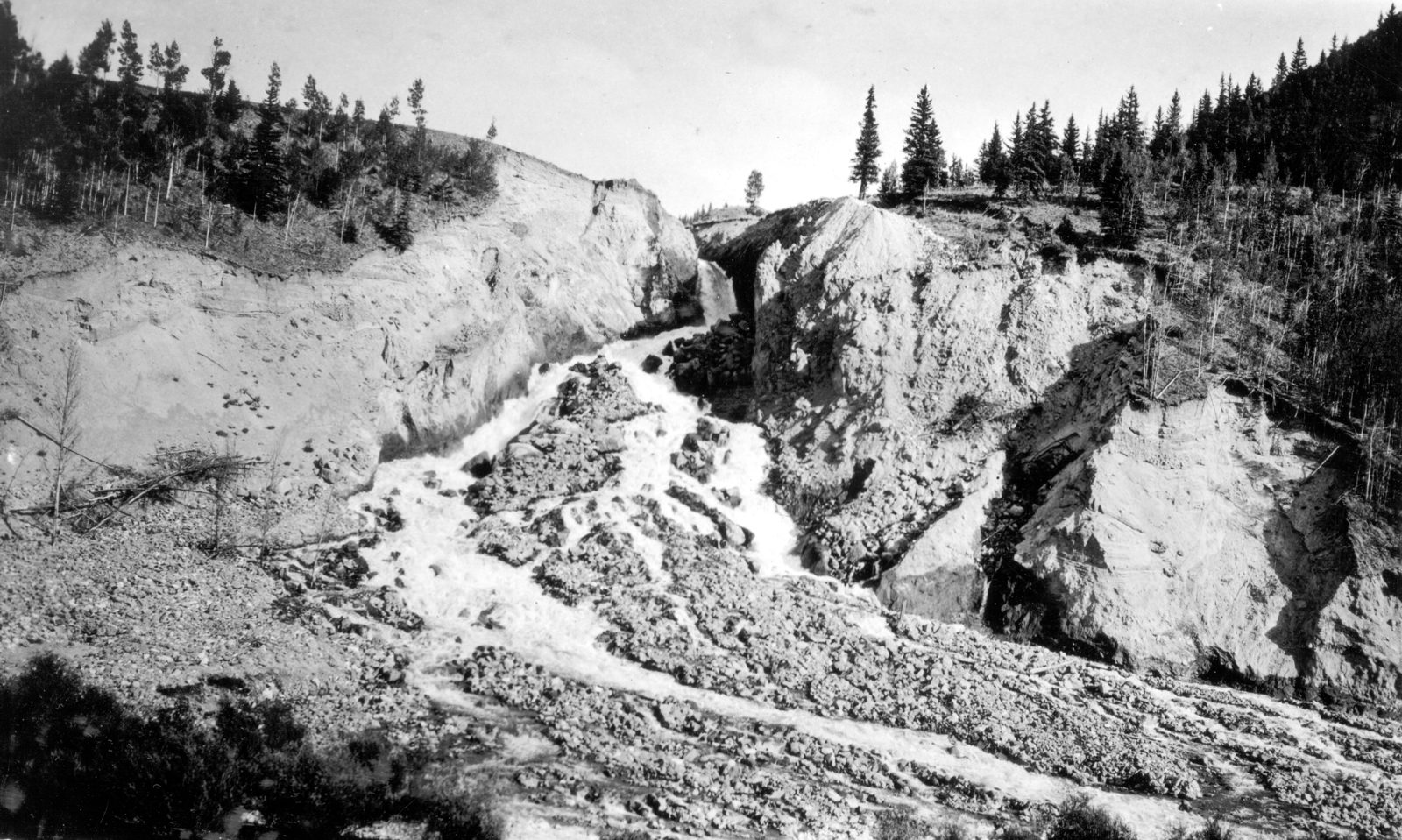 A rough channel cut through pale broken ground below a dam, scattered timber and rubble along its sides, with conifers on the rim above.
